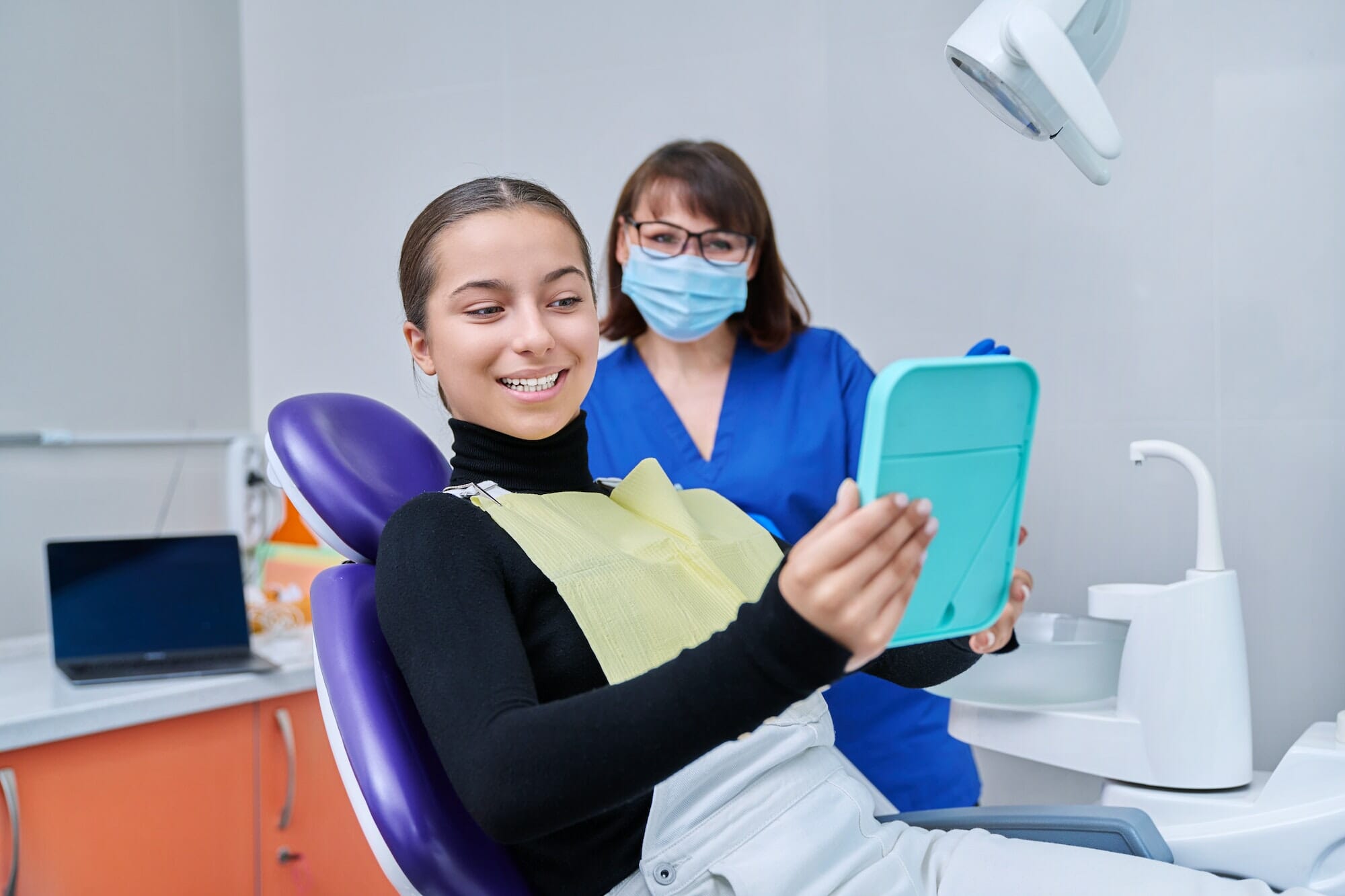 Happy female with mirror sitting in dentist chair looking at healthy teeth